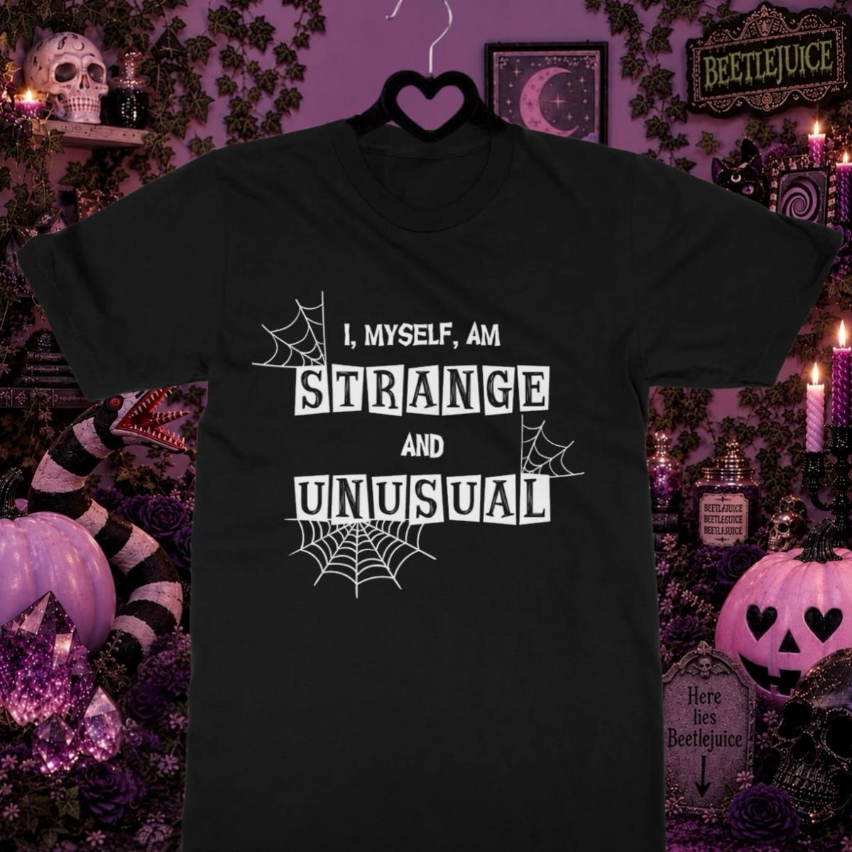 A black short-sleeve Beetlejuice inspired t-shirt hanging on a black hanger against a Halloween-themed background. The shirt features a white text graphic that reads 'I, MYSELF, AM STRANGE AND UNUSUAL' with spiderweb illustrations.
