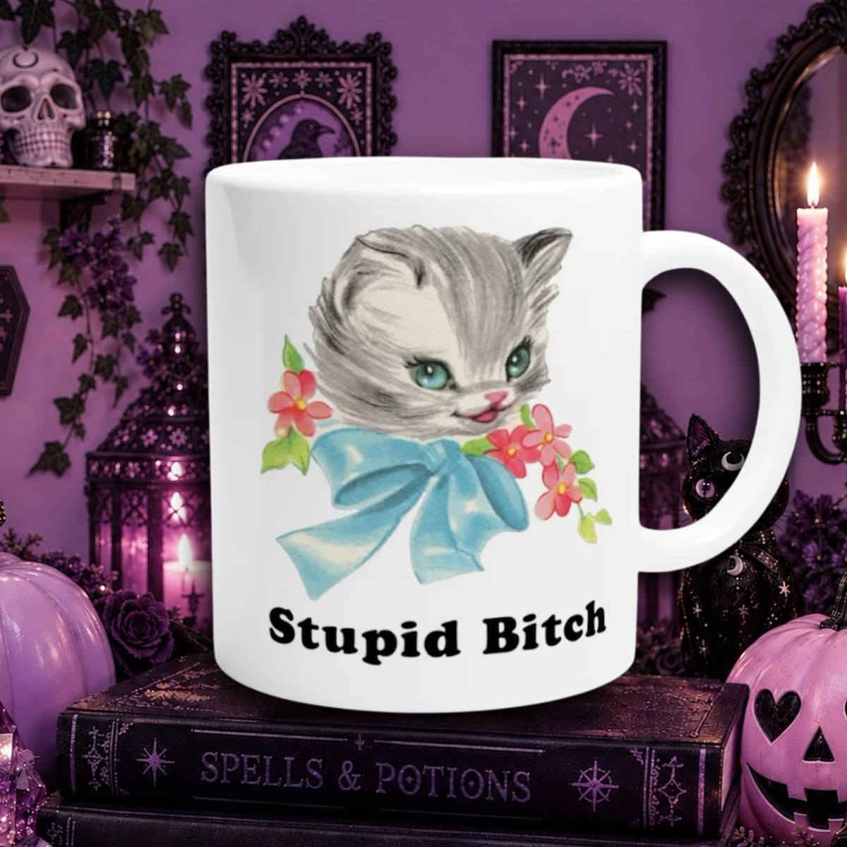 A white ceramic mug featuring a vintage-style illustration of a cute cat, surrounded by pink flowers, with the text 'Stupid Bitch' printed below. The mug is displayed on a dark book in a gothic-themed setting with purple walls, candles, and decorative Halloween props.