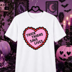 A white short-sleeve t-shirt on a black hanger, featuring a pink heart graphic with a red scalloped border and the text 'THICK THIGHS SAVE LIVES' in black. The shirt is displayed against a dark purple Halloween-themed background with pumpkins, candles, and gothic decor.