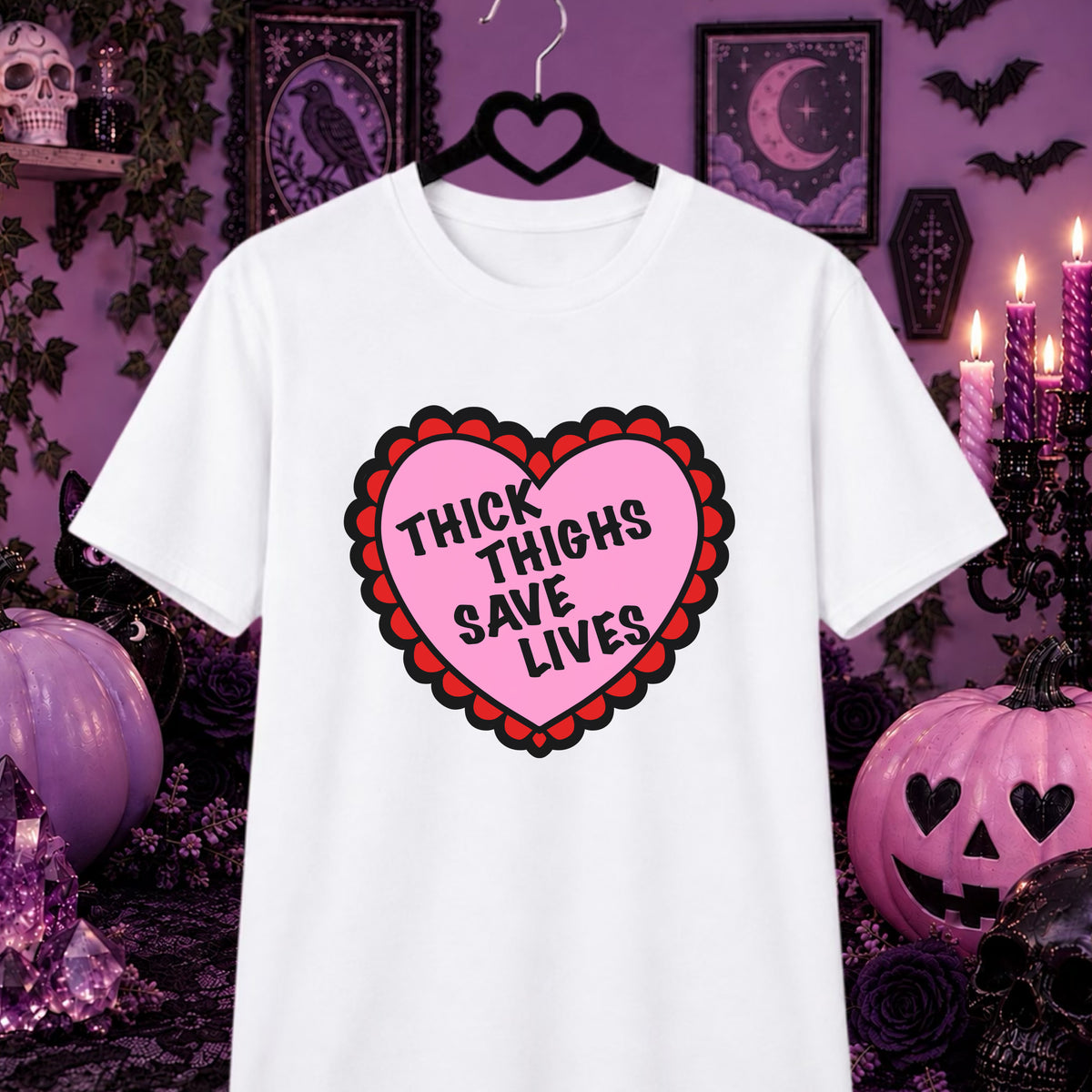 A white short-sleeve t-shirt on a black hanger, featuring a pink heart graphic with a red scalloped border and the text 'THICK THIGHS SAVE LIVES' in black. The shirt is displayed against a dark purple Halloween-themed background with pumpkins, candles, and gothic decor.