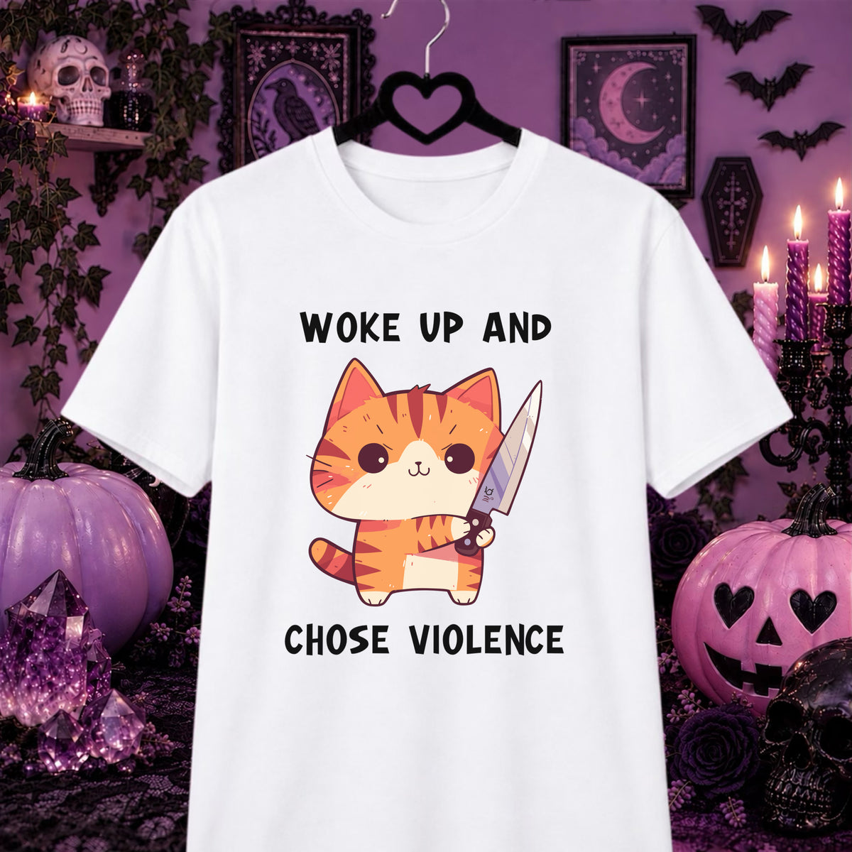 A white short-sleeve graphic T-shirt hanging on a black hanger against a Halloween-themed purple background. The shirt features a cartoon orange tabby cat holding a knife with the text 'WOKE UP AND CHOSE VIOLENCE' in black lettering.